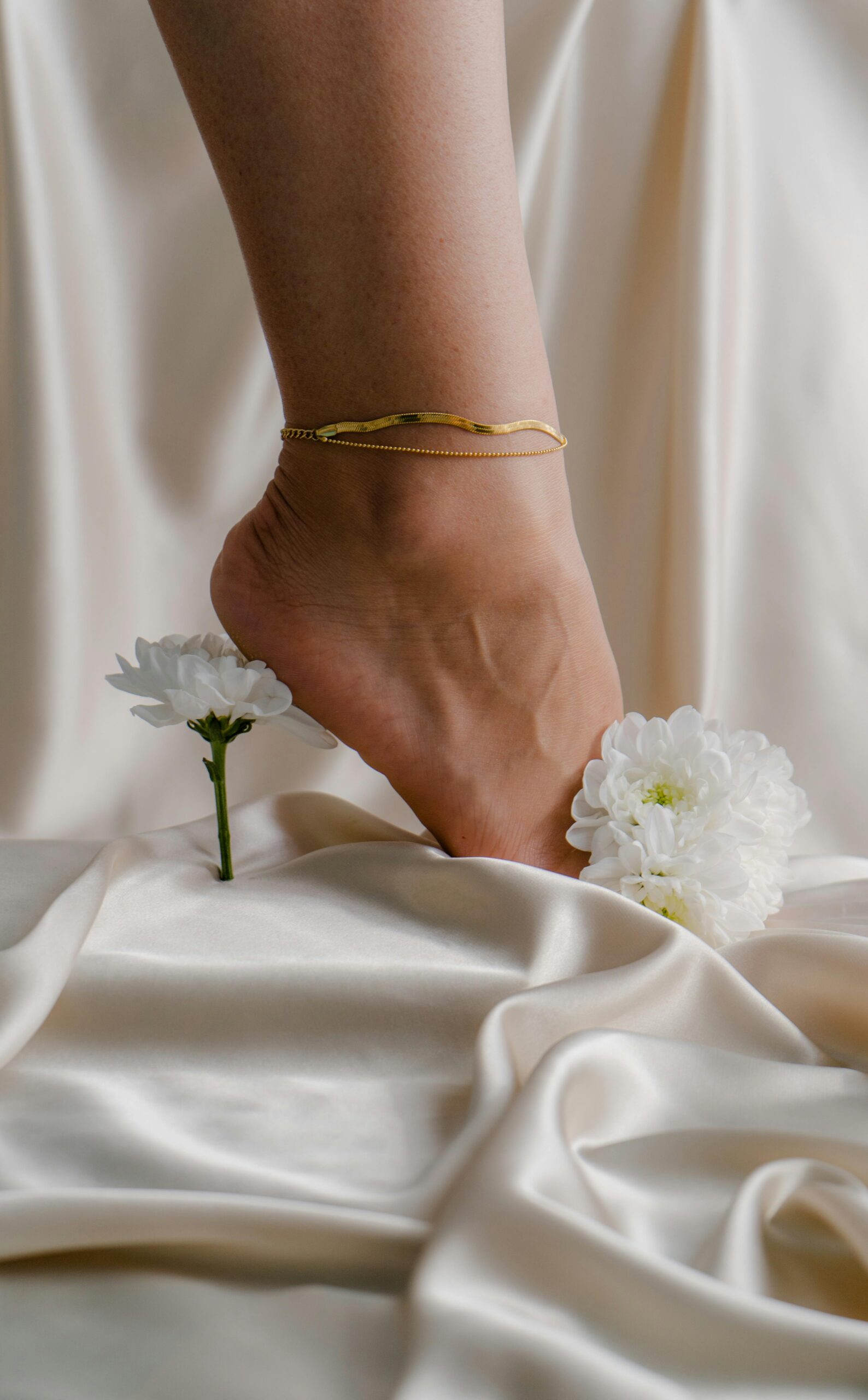 A graceful foot with gold anklet on luxurious silk, surrounded by white flowers.